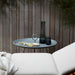 Gray metal round outdoor tray table with a transparent black bottle and glass on top, placed against a wooden fence