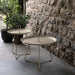Two beige round metal outdoor tray tables in different sizes, each with a circular center handle, placed on a patio against a stone wall
