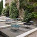 A series of modern circular outdoor tray tables in various colors, set against a stone wall and lush greenery