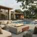 Modern outdoor patio scene with a rectangular concrete fire pit table burning, surrounded by cream outdoor seating, a patterned rug, a swimming pool, and a contemporary stone-and-wood house with trees in the background.