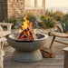 A round concrete fire pit bowl with a pedestal base, filled with burning wood and flames on a wooden deck. A beach house, plants, and outdoor chairs are visible in the background, creating a cozy patio atmosphere.