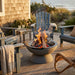 A round concrete fire pit with an active fire in the center, set on a wooden deck surrounded by blue Adirondack chairs and a shingled house exterior at sunset.