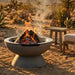 A round concrete fire pit with a pedestal base, shown outdoors in a desert setting with a fire burning inside and wooden logs on fire. A small wooden stool and part of a cushioned chair are visible in the background.