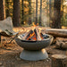 A round concrete fire pit bowl with a pedestal base, holding burning logs and embers outdoors in a forest clearing. A folding camp chair, log bench, and bag are visible in the softly blurred background.