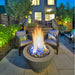 Modern patio with gray fire pit bowl, two gray lounge chairs, and garden setting at night.