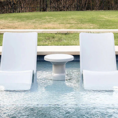 Two modern white in-pool chaise loungers  and a round side table on a pool sunshelf with green lawn in the background.