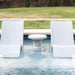 Two modern white in-pool chaise loungers  and a round side table on a pool sunshelf with green lawn in the background.