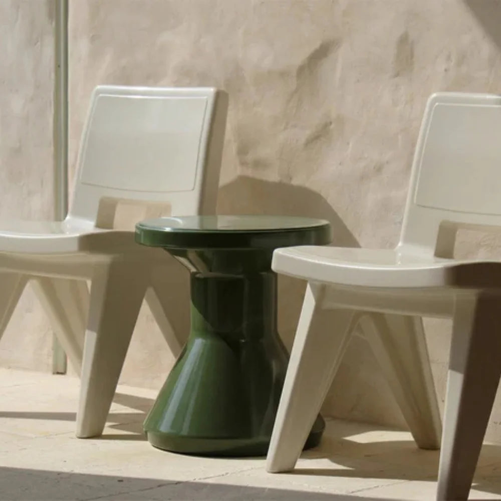 Two white chairs with a small green round outdoor side table between them against a beige wall.