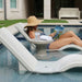 Two light gray in-pool chaise loungers and a round side table, with a woman in a sun hat relaxing on one lounger.