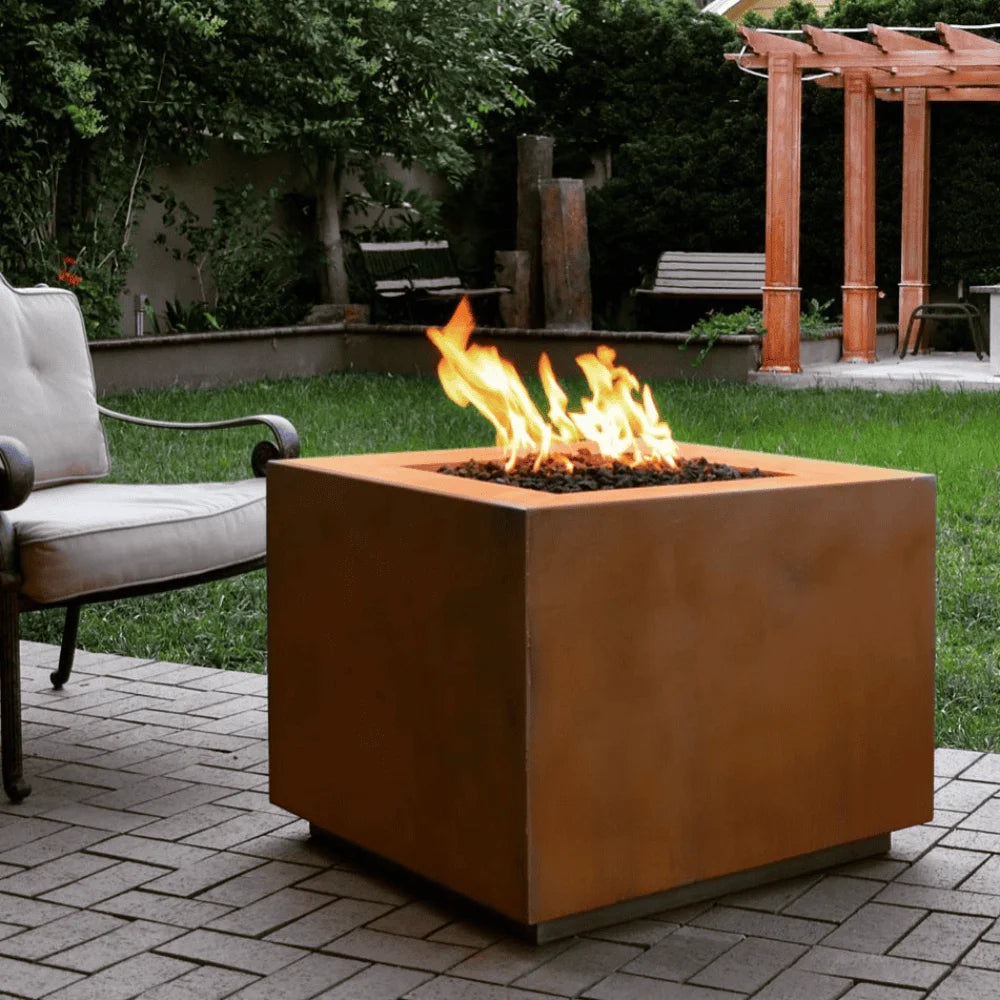A cube shaped corten steel outdoor square metal fire pit with flame set on a paved patio area beside cushioned outdoor chair