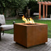 A cube shaped corten steel outdoor square metal fire pit with flame set on a paved patio area beside cushioned outdoor chair