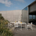 Rectangular white outdoor dining table paired with four white dining chairs, set on a wooden deck patio with tall grasses.