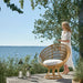 Natural rattan outdoor sun chair with a white cushion beside a person on a seaside patio.