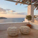 Natural wicker outdoor ottomans in a light tan finish with open woven frames on a seaside terrace.