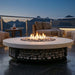 Round outdoor fire pit table with a light stone top, black metal base filled with stones, and active flames, set on a patio at dusk with lounge chairs and a railing in the background.