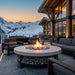 A round outdoor fire pit table with a stone base and glowing flames, surrounded by modern gray patio furniture on a deck overlooking a snowy mountain landscape at sunset.