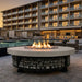 A round outdoor fire pit with a light beige stone top and a black metal base filled with gray stones, burning with bright orange flames. It is situated on a stone patio next to a swimming pool, with a modern multi-story hotel building in the background at dusk.
