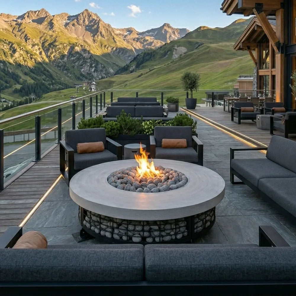 Outdoor round fire pit table with a glowing fire, set on a modern patio with dark gray lounge chairs and sofas, overlooking a scenic mountain landscape.