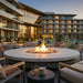 A round outdoor fire pit table with a glowing fire, set in a modern patio lounge area at dusk with resort-style buildings and a pool in the background.
