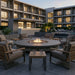 Outdoor patio furniture set with a round stone fire pit surrounded by cushioned wooden chairs and a small matching side table, set in a modern resort-style poolside lounge area at dusk.