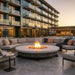 A round outdoor fire pit with a glowing fire, surrounded by a curved sectional sofa on a patio in front of a modern high-rise building at sunset.
