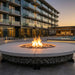 A round outdoor fire pit with a glowing fire in the center, surrounded by small gray stones and a light concrete top, set in a modern patio area with a pool and contemporary apartment buildings in the background at dusk.