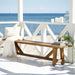 Natural wooden outdoor bench with a white pillow and blanket beside books and candles on a seaside porch.