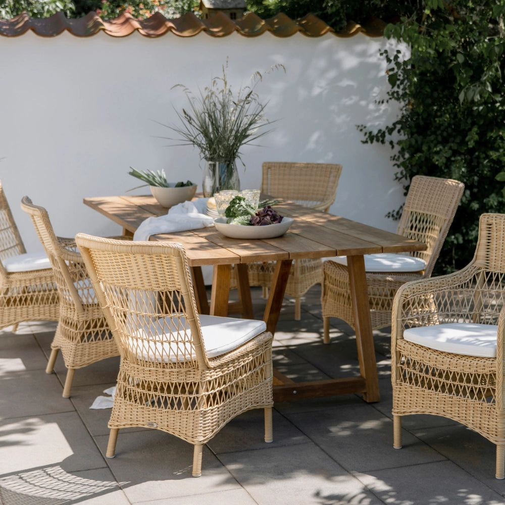 Natural wooden outdoor dining table with six matching wicker dining chairs in a garden setting.