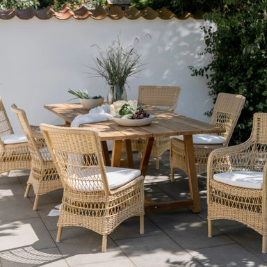 Natural wooden outdoor dining table with six matching wicker dining chairs in a garden setting.