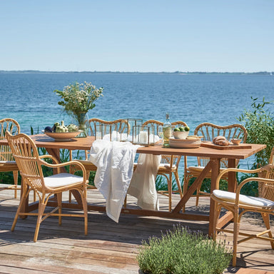 Natural wooden outdoor dining table with matching wicker dining chairs in a seaside setting.