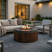 A round outdoor fire pit table with a dark wood slatted base and a concrete top, burning with a visible flame in a modern patio setting with a sofa and lounge chairs.