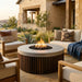 A round outdoor fire pit table with a concrete top and dark wood slatted base, lit with a visible flame, placed in a patio seating area with upholstered chairs, a sofa, pillows, plants, and a stucco house in the background.