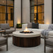 Outdoor patio with round concrete outdoor fire pit table, chairs, and a couch in front of a building with large windows.