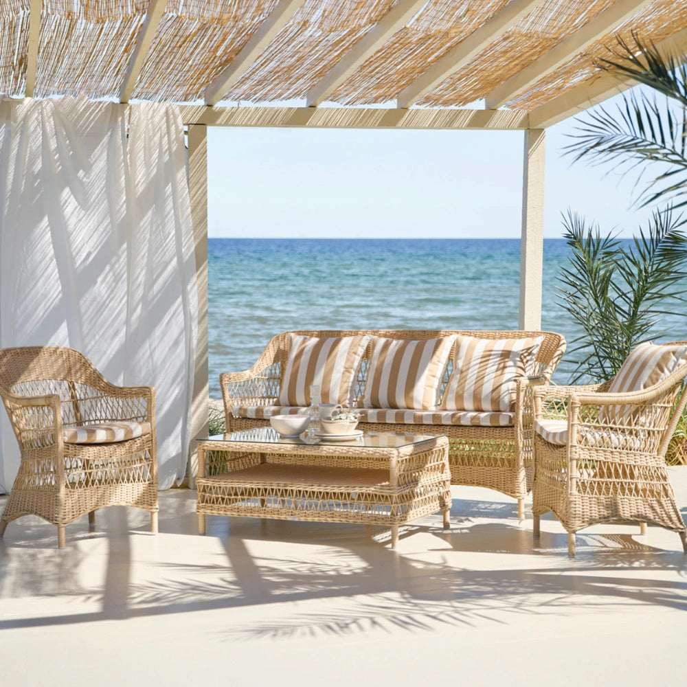 Natural wicker outdoor coffee table with a glass top and matching sofa and lounge chairs featuring striped cushions.