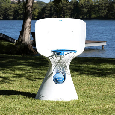 A white outdoor basketball hoop with a blue rim and net stands on a grassy lawn near a lake, with a tree and dock visible in the background. A blue basketball is hanging from the hoop.