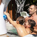 A man and two young boys play with a blue SLINKS basketball in a pool. A white outdoor basketball hoop and net are attached to a white wall, with patio furniture and trees in the background.