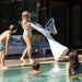 A woman in a light blue swimsuit walks beside a swimming pool while holding a large white slide with a blue basketball hoop attached. Two children are standing by the pool, and another child is swimming in the water.