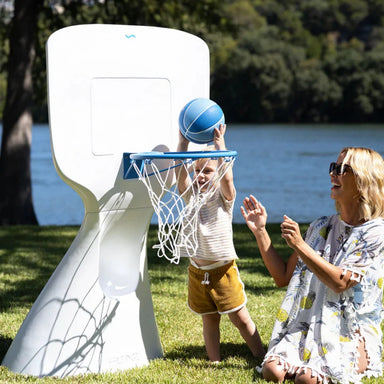A child throws a blue basketball into a white and blue outdoor hoop while a woman watches from beside it on a grassy lawn with water and trees in the background.