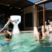 A white pool-side basketball hoop with a curved backboard stands in a swimming pool, with several people playing and relaxing around it in an outdoor patio setting.