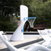 A white outdoor basketball hoop stand with a blue rim and white net, positioned beside a pool with lounge chairs and blurred greenery in the background.