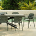 Gray rectangular concrete outdoor dining table with black angular metal legs and five green chairs beside pool with trees in background