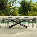 Modern rectangular concrete outdoor dining table with six green chairs placed next to a pool, surrounded by lush green trees