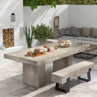 White patio with rectangular concrete dining table and concrete outdoor bench with black metal legs, plants and corner seating with gray cushions and throw pillows