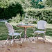 White rattan outdoor dining armchairs with a matching round wooden dining table in a lush garden.