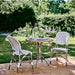 Natural rattan outdoor dining side chairs beside a matching round wooden bistro table on a brick patio.