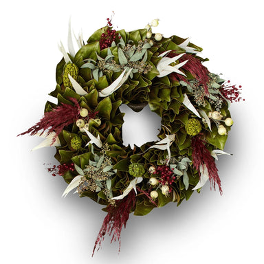 Festive wreath with green magnolia leaves, white leaves, red accents, and delicate dried flowers on a white background.