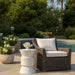 White speckled concrete side table beside wicker chair with white cushions and potted plants near a pool, surrounded by greenery