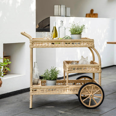 Natural wicker outdoor bar cart with rear wheels and two shelves styled with bottles and glassware.