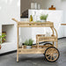 Natural wicker outdoor bar cart with rear wheels and two shelves styled with bottles and glassware.
