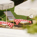 Natural rattan outdoor sun lounger with white cushions, rear wheels and red striped towel on a sunny patio.